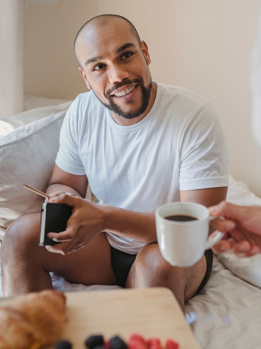 Homem sorrindo de camiseta branca e cueca boxer preta no cafe da manha